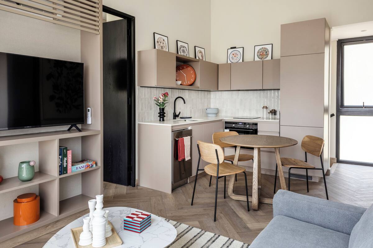 Un salon moderne avec un téléviseur, un coin repas avec une table et des chaises, une cuisine équipée de placards et d’une horloge, et un canapé.