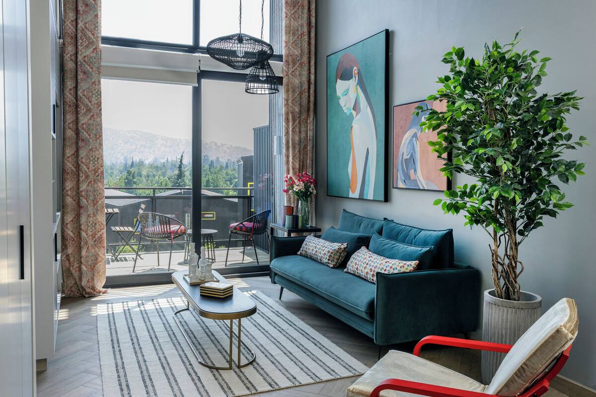Un salon moderne avec un canapé, un fauteuil rouge, une plante en pot et une vue sur la verdure grâce à une grande fenêtre.