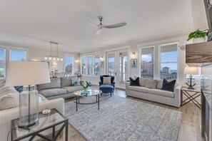 Ein geräumiges Wohnzimmer mit modernem Design, ausgestattet mit Deckenventilator, großen Fenstern und einer Auswahl an Sitzmöglichkeiten, darunter ein Sofa, ein Sessel und ein Couchtisch.