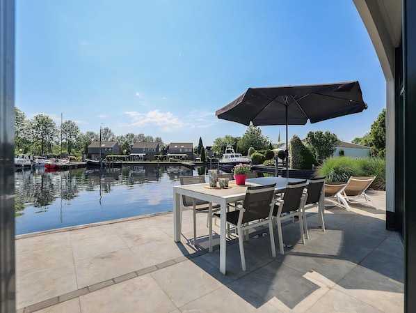 Un patio con mesa y sillas, un gran paraguas y vistas a un puerto deportivo con barcos y casas.