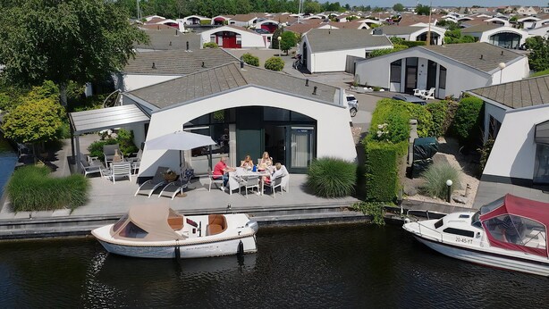 A modern waterfront property with a covered outdoor seating area, a boat docked nearby, and a row of similar houses in the background. - Suny bungalow with a private jetty. (Lemmer)