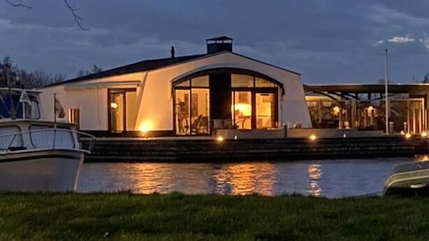 Ein modernes Hausboot mit großem Panoramafenster, überdachter Terrasse und Anlegesteg.