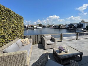 Un patio frente al agua con mobiliario de mimbre y vistas a barcos y edificios al otro lado.