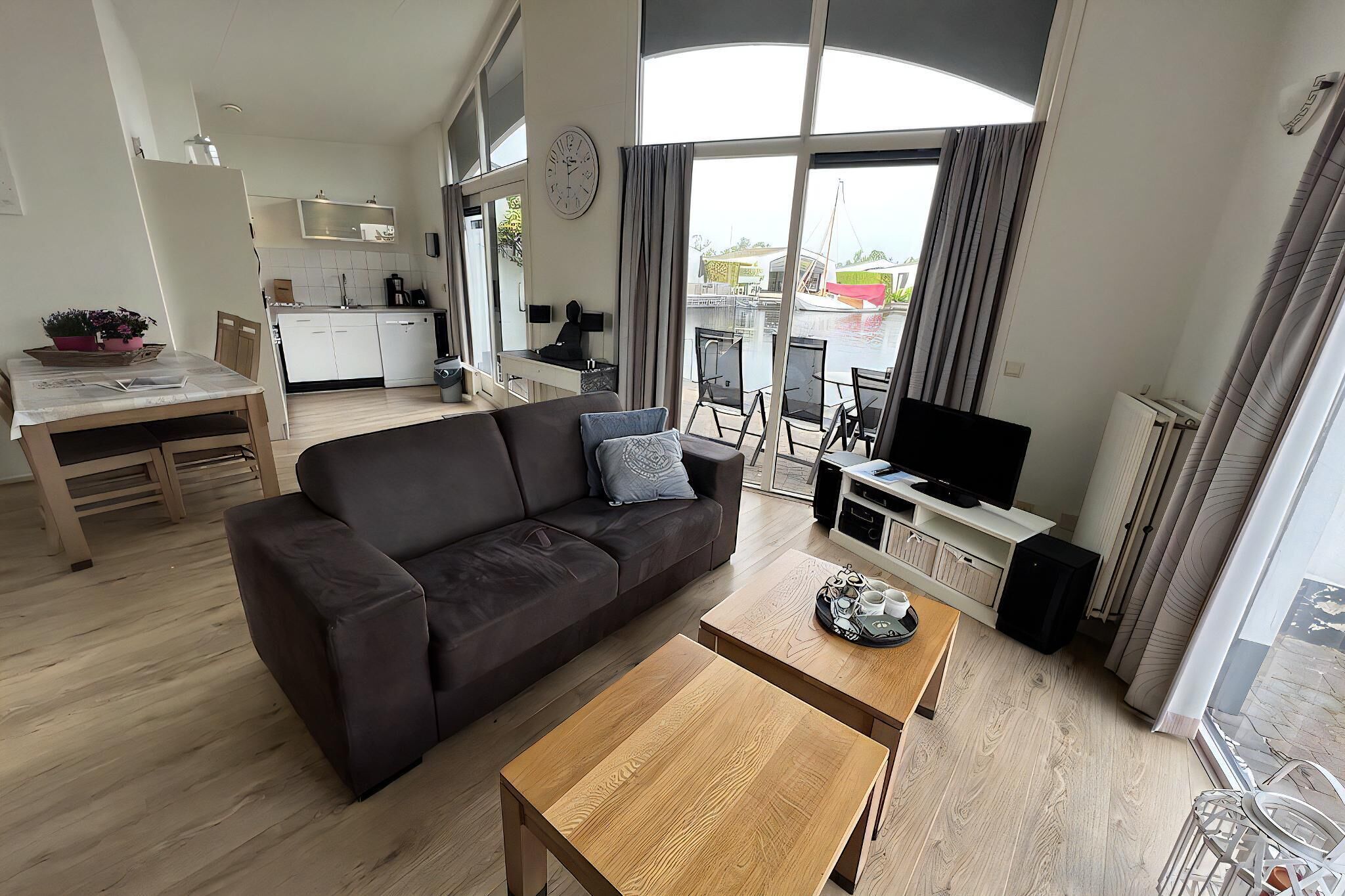Una sala moderna con sofá, mesa de centro y televisor. Se aprecia en el fondo una cocina con gabinetes blancos y un comedor con mesa y sillas.
