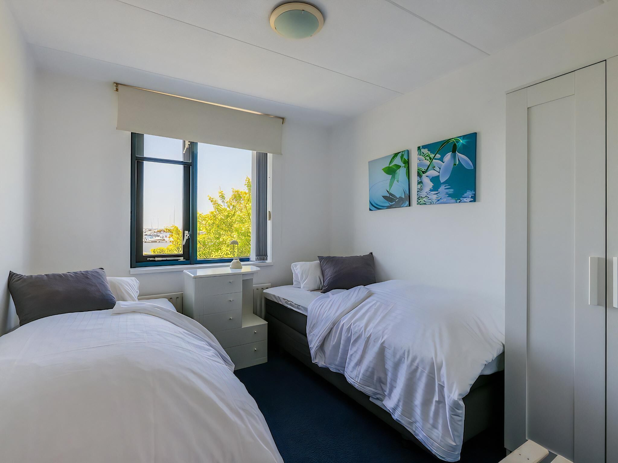 A bedroom with two beds, a desk, and a window with a view of trees and a building.