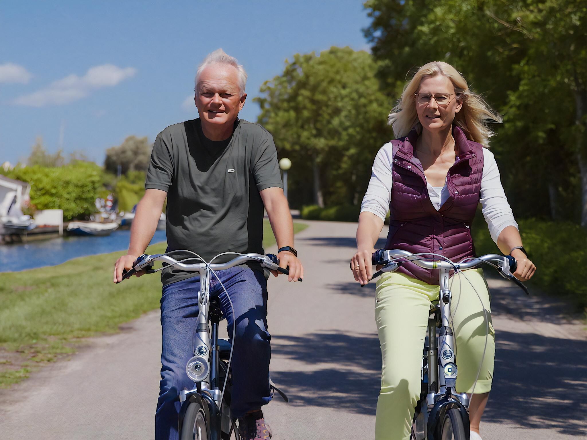 Two people riding bicycles on a paved path with greenery and a body of water in the background.