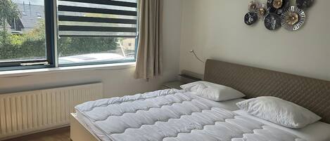 Um quarto com uma cama, uma janela com persianas, um radiador e detalhes decorativos nas paredes.