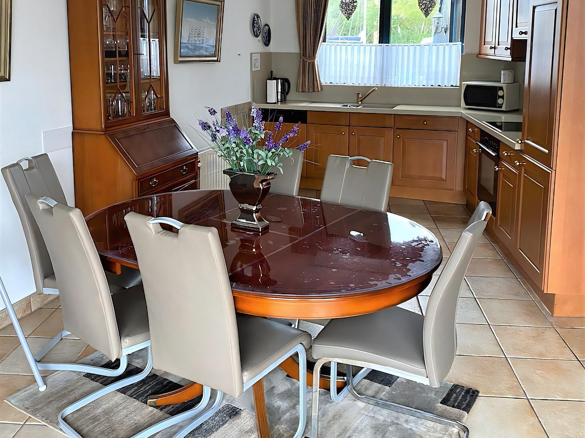 Ein Essbereich mit einem runden Tisch, sechs Stühlen, einer Blumenvase und einer Küche im Hintergrund.