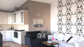 Een moderne woonkamer met een zwarte bank, een witte bank en een salontafel. Er is een keuken met witte kasten en een eettafel.