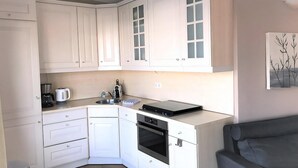 Een kleine keuken met witte kasten, een inbouwoven, een koffiezetapparaat en een eethoek met een grijze bank.