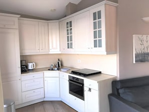 Een kleine keuken met witte kasten, een inbouwoven, een koffiezetapparaat en een eethoek met een grijze bank.