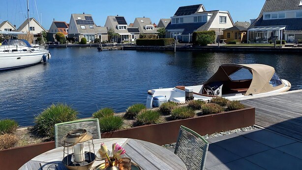 Een jachthaven met aangemeerde boten, een houten terras met buitenmeubilair en huizen op de achtergrond.