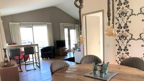 Een moderne woonkamer met een houten eettafel, comfortabele zithoek, een flatscreen televisie en een groot raam met gordijnen.