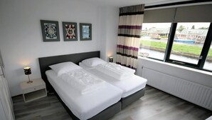 A modern hotel room with a large bed, a nightstand, and a window with a view of a building and greenery. - Park Brekkense Wiel 038 (Lemmer)