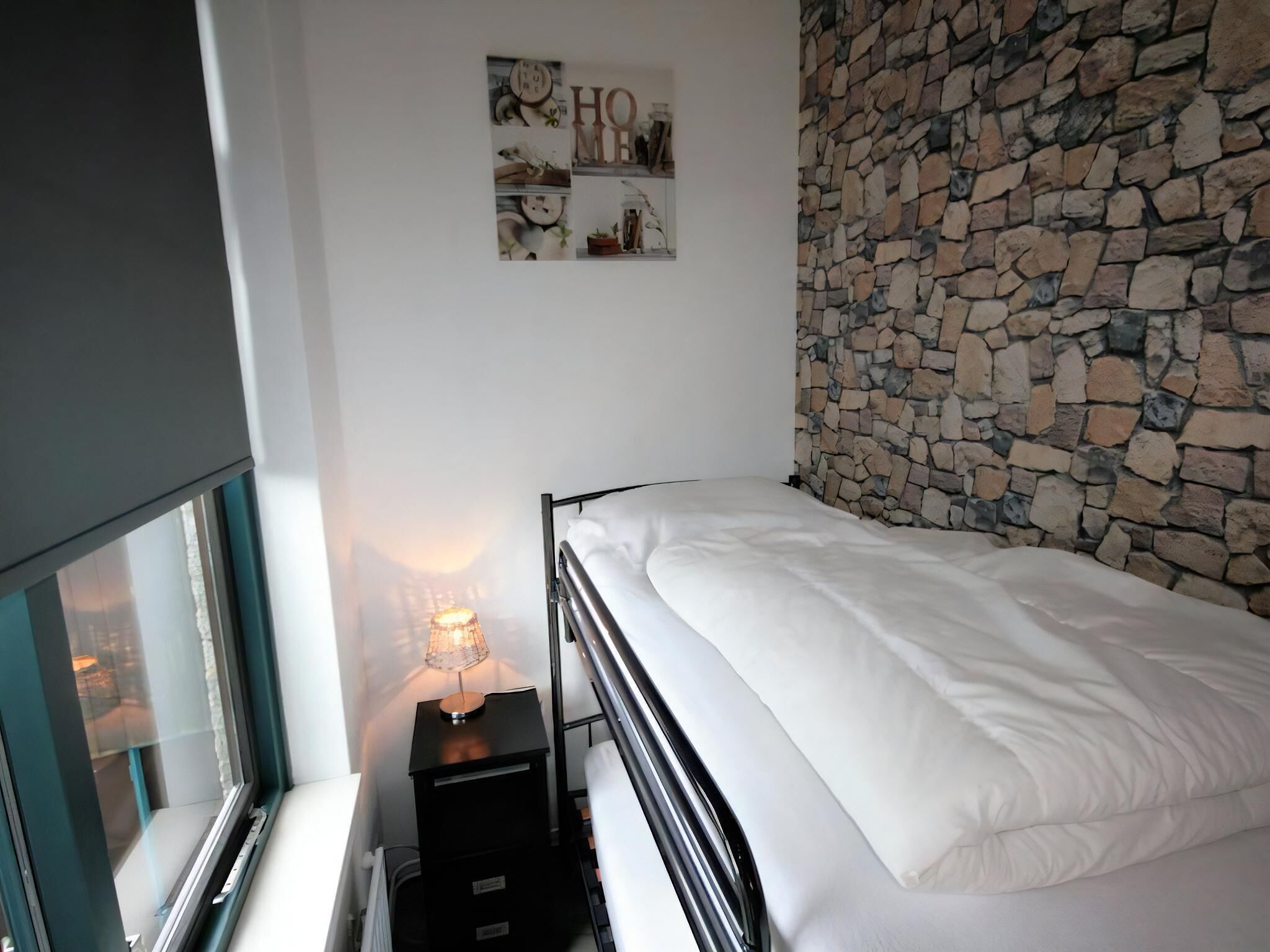A bedroom with a stone wall, a bed, a bedside table with a lamp, and a window.