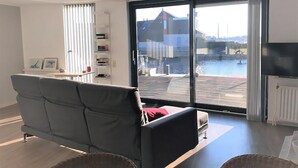 A modern living room with a grey sofa, a small table, and a large window offering a view of a dock and water. - Park Brekkense Wiel 229 (Lemmer)