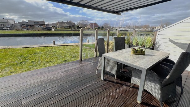 Une terrasse extérieure couverte, dotée d’une table et de chaises, qui offre une vue sur un canal, avec des bateaux et des maisons en arrière-plan.