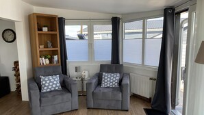 A modern living room with two grey armchairs, a wooden bookshelf, and a view of the outside through large windows.