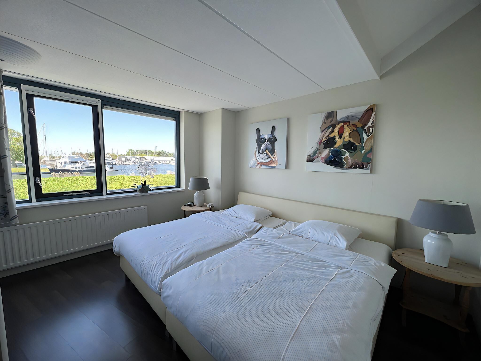 Ett hotellrum med en stor säng, två sängbord, ett fönster med utsikt över båtar och två tavlor som hänger på väggen.