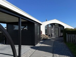 Ein modernes Gebäude mit überdachter Terrasse, einem Weg zum Eingang und einer kleinen Sitzgelegenheit mit Stühlen.