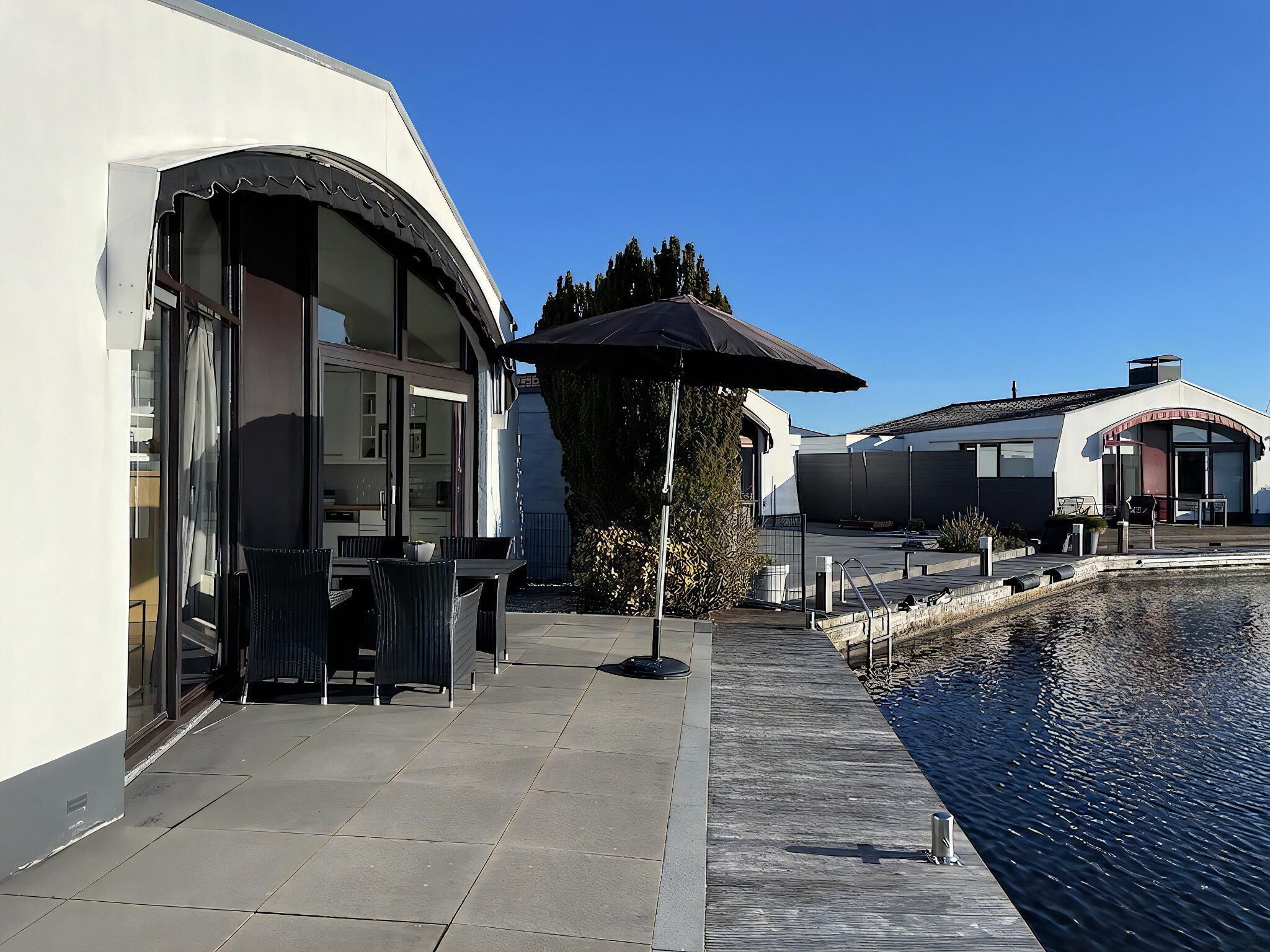 A modern outdoor patio with a pool, a dining area with chairs and a table, and a covered seating area.