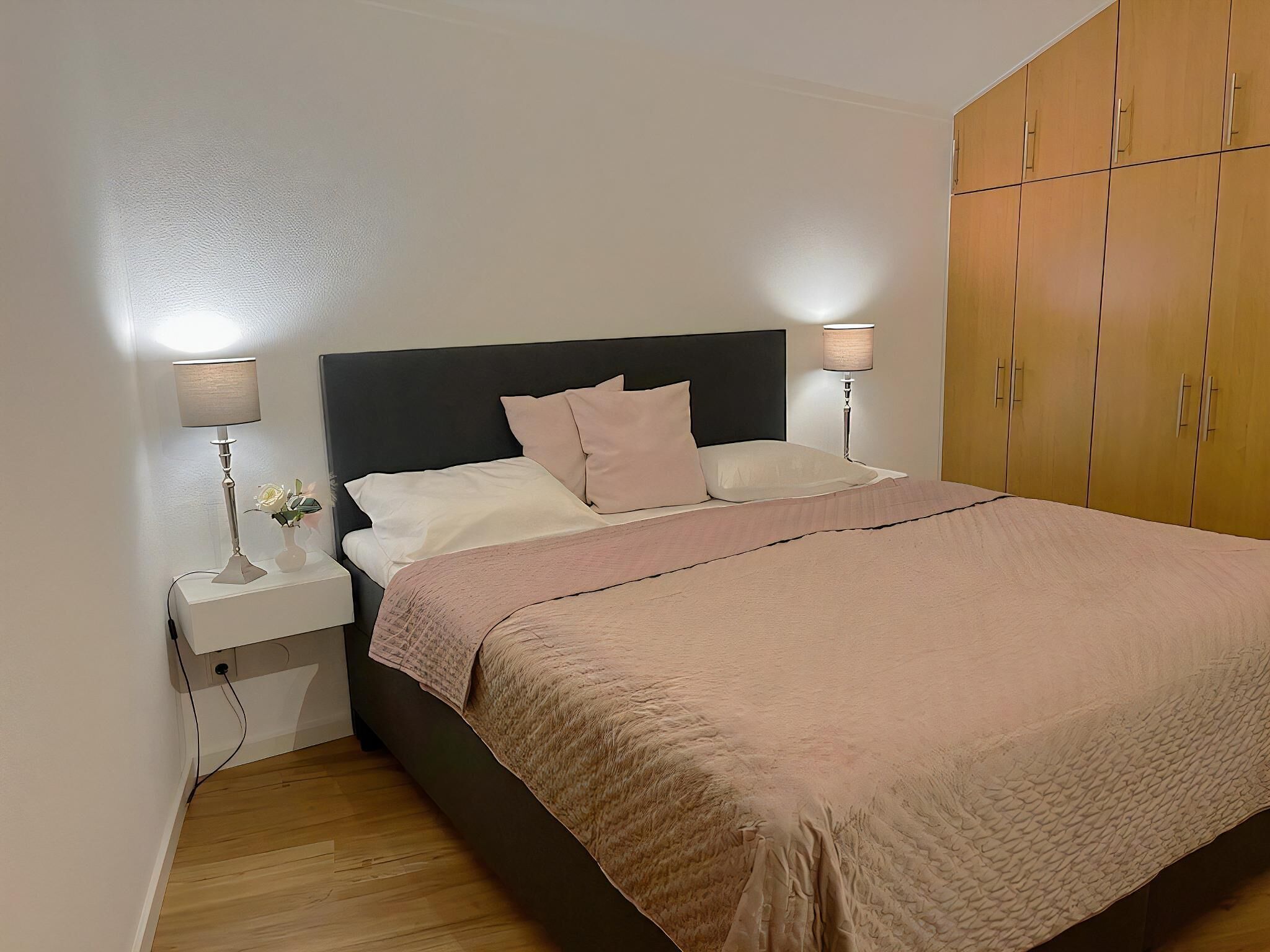 Een opgemaakt bed met een donker hoofdbord, twee bedlampjes en een houten kledingkast.