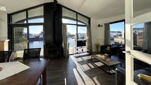 Un salon moderne doté d’une cheminée, d’un coin repas et de grandes fenêtres offrant une vue sur un plan d’eau et des bâtiments.