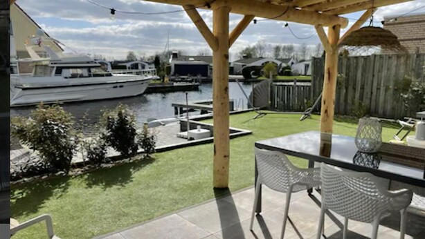 Une terrasse couverte avec une table et des chaises, un petit jardin et une vue sur une marina remplie de bateaux.
