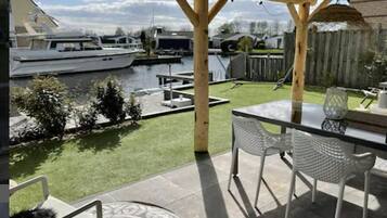 Een overdekt terras met een tafel en stoelen, een kleine tuin en uitzicht op een jachthaven met boten.