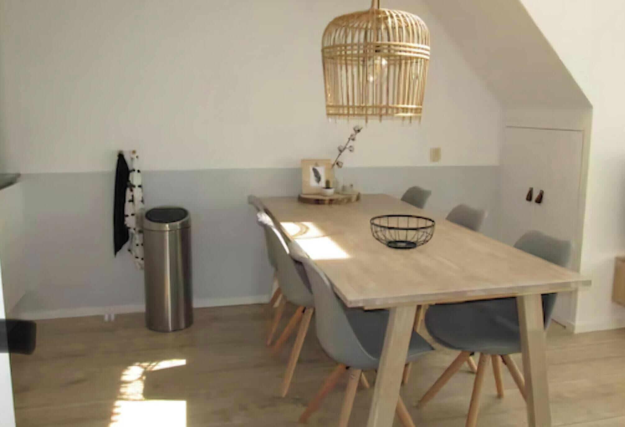 Een eetgedeelte met een lange houten tafel, grijze stoelen, een rieten hanglamp en een kleine prullenbak.