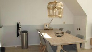 Un coin repas avec une longue table en bois, des chaises grises, un luminaire suspendu en osier et une petite poubelle.