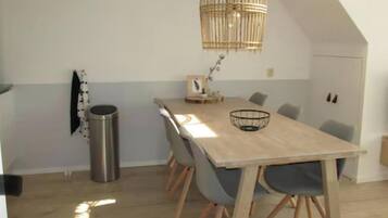 Een eetgedeelte met een lange houten tafel, grijze stoelen, een rieten hanglamp en een kleine prullenbak.