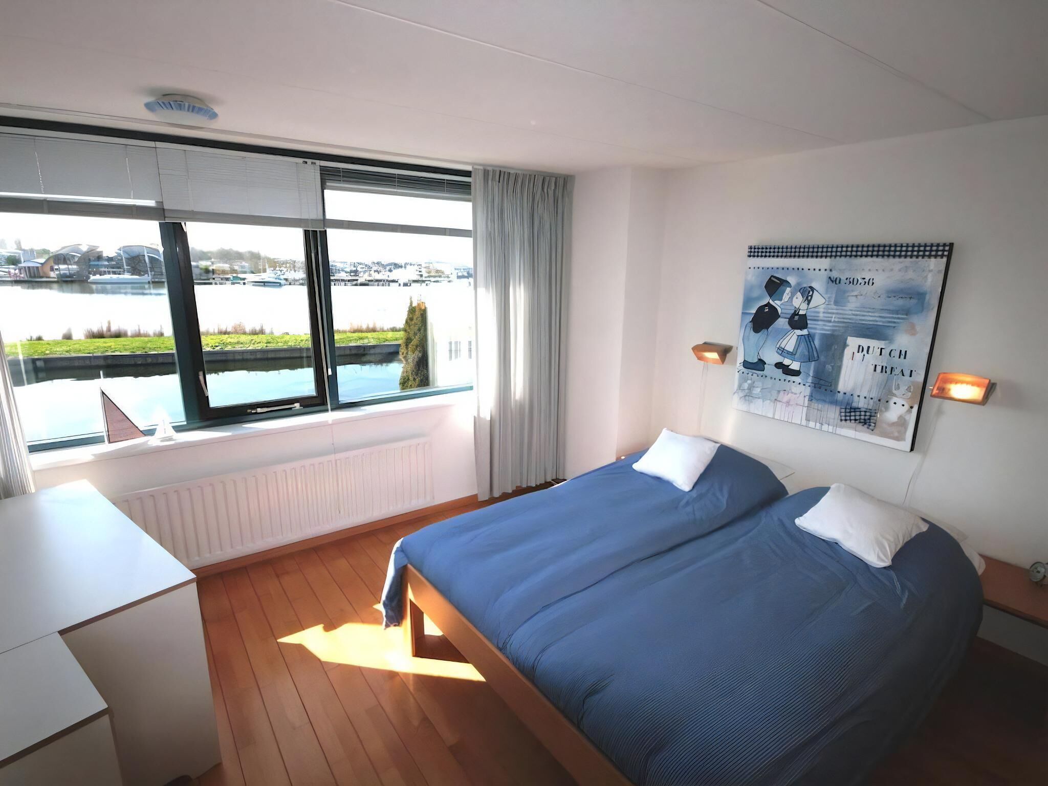 A hotel room with a bed, a desk, a window with a view of a body of water, and a wall-mounted artwork.