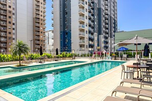 A swimming pool with lounge chairs and umbrellas, surrounded by high-rise residential buildings. - Moderno com AC piscina e academia (Rio de Janeiro)
