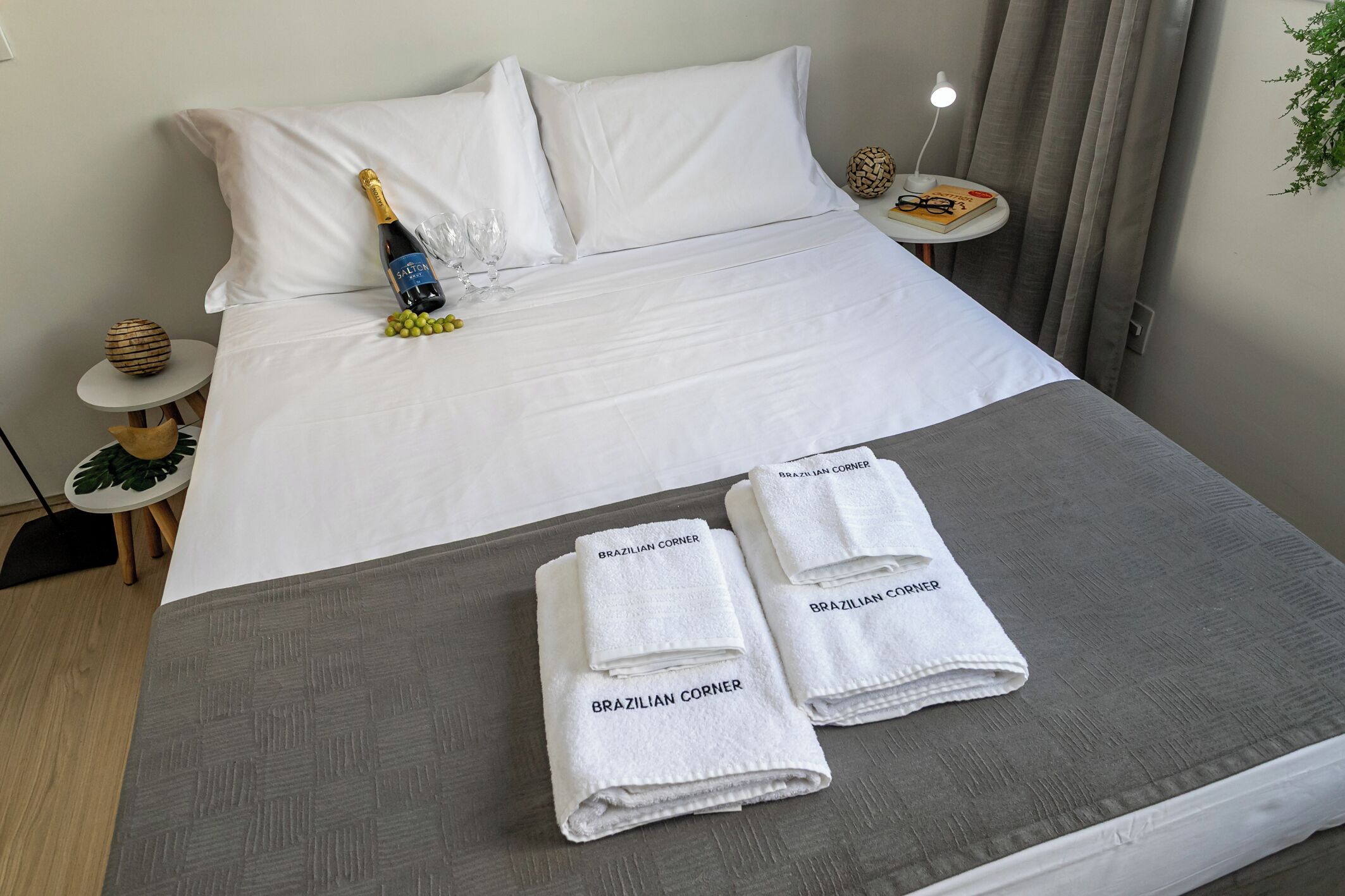 Una cama bien hecha con sábanas blancas, una botella de champán y dos toallas con la marca 'Brazilian Corner'.