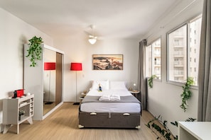 Quarto de hotel com cama, mesa de cabeceira, abajur, espelho, janela e vista para prédios.