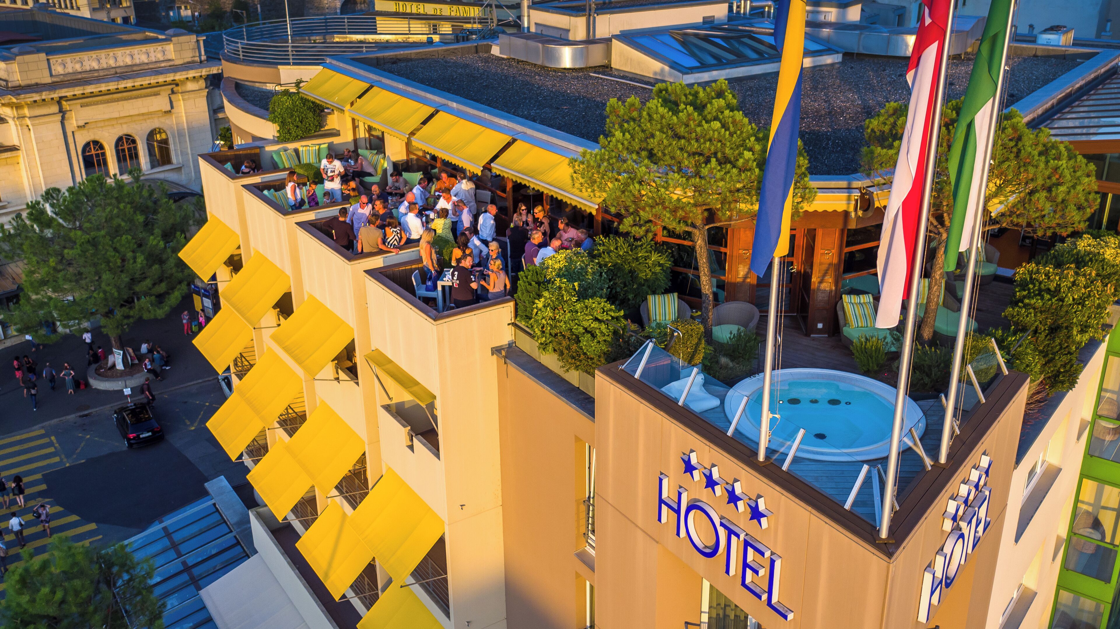 Ein Hotel mit Dachterrasse, Bar und Poolbereich, umgeben von viel Grün und Flaggen.