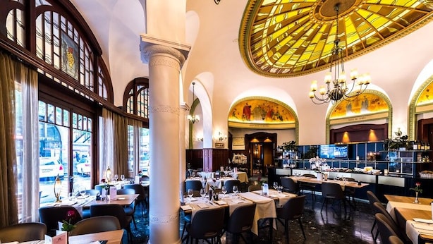 A restaurant with high ceilings, arched windows, and a central chandelier. Tables are set with white tablecloths and wine glasses. - Astra Studio (Vevey)