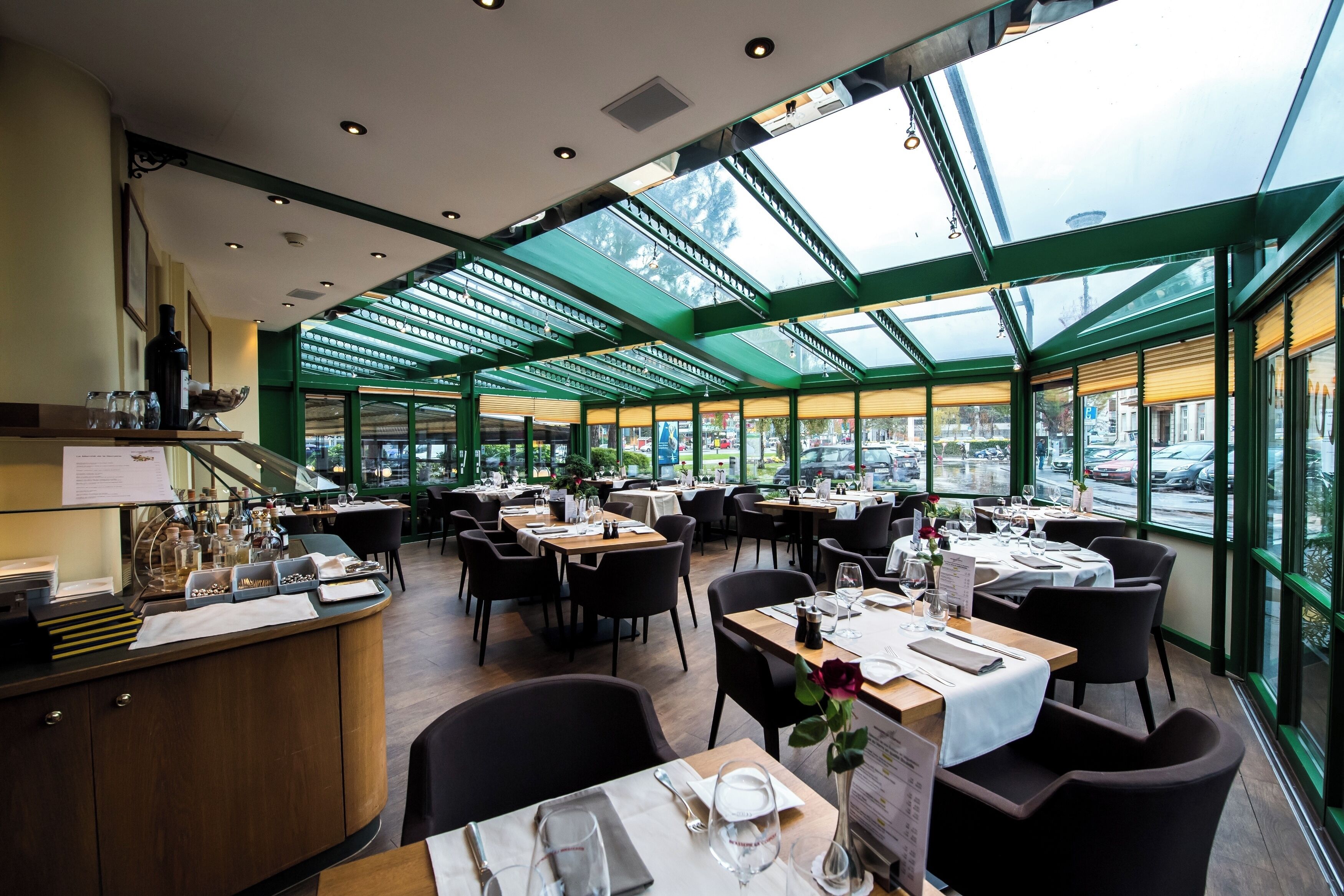 En restaurant med et glasindrammet spiseområde, flere borde dækket til spisning, og udsigt til gaden.