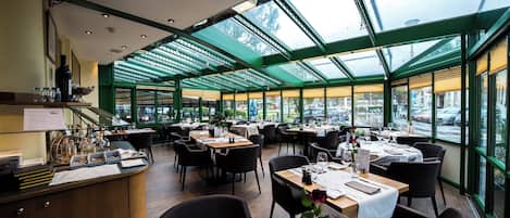 En restaurant med et glasindrammet spiseområde, flere borde dækket til spisning, og udsigt til gaden.