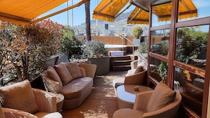 A sunlit outdoor seating area with wicker furniture and a wooden deck, overlooking a mountainous landscape. - Astra Studio (Vevey)