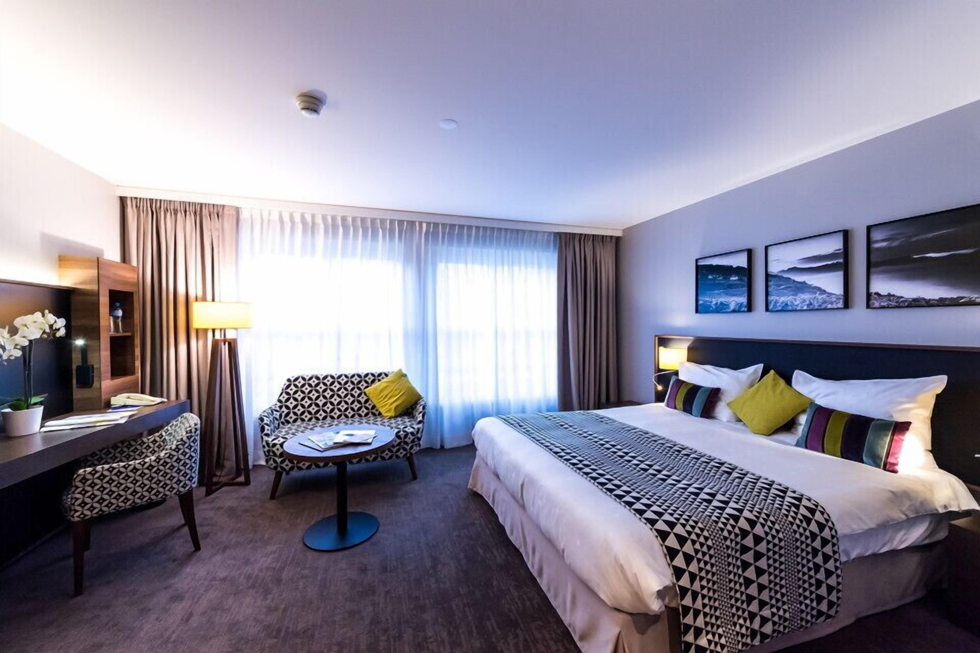 Une chambre d’hôtel avec un lit, un bureau, une chaise et une petite table. Il y a des rideaux, une lampe et trois tableaux encadrés au mur.
