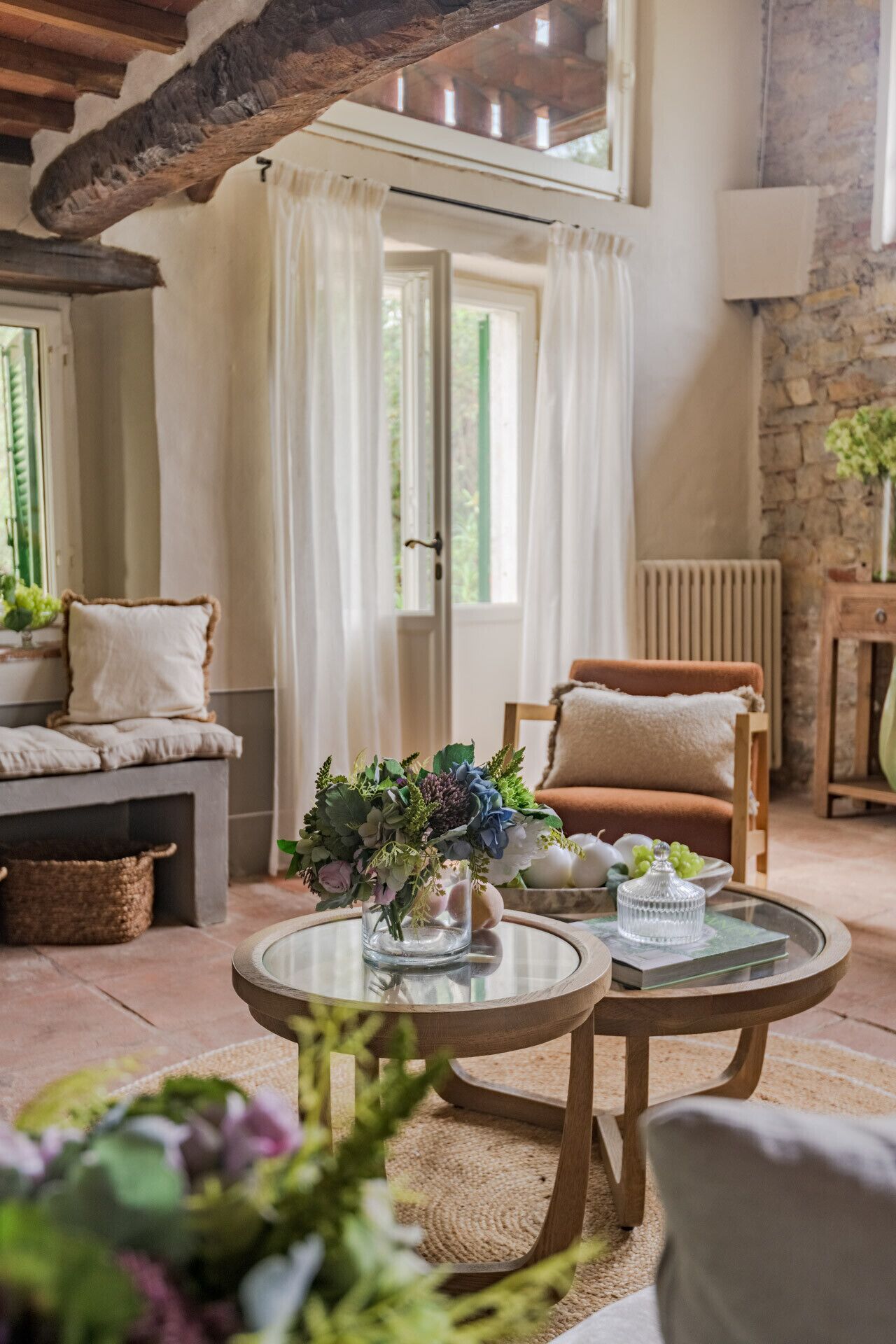 A cozy living room with a stone wall, a wooden ceiling, a sofa with cushions, a chair, and a glass table with a vase of flowers.