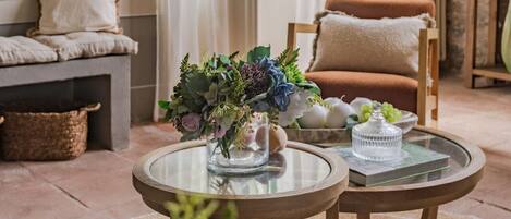 A cozy living room with a stone wall, a wooden ceiling, a sofa with cushions, a chair, and a glass table with a vase of flowers.