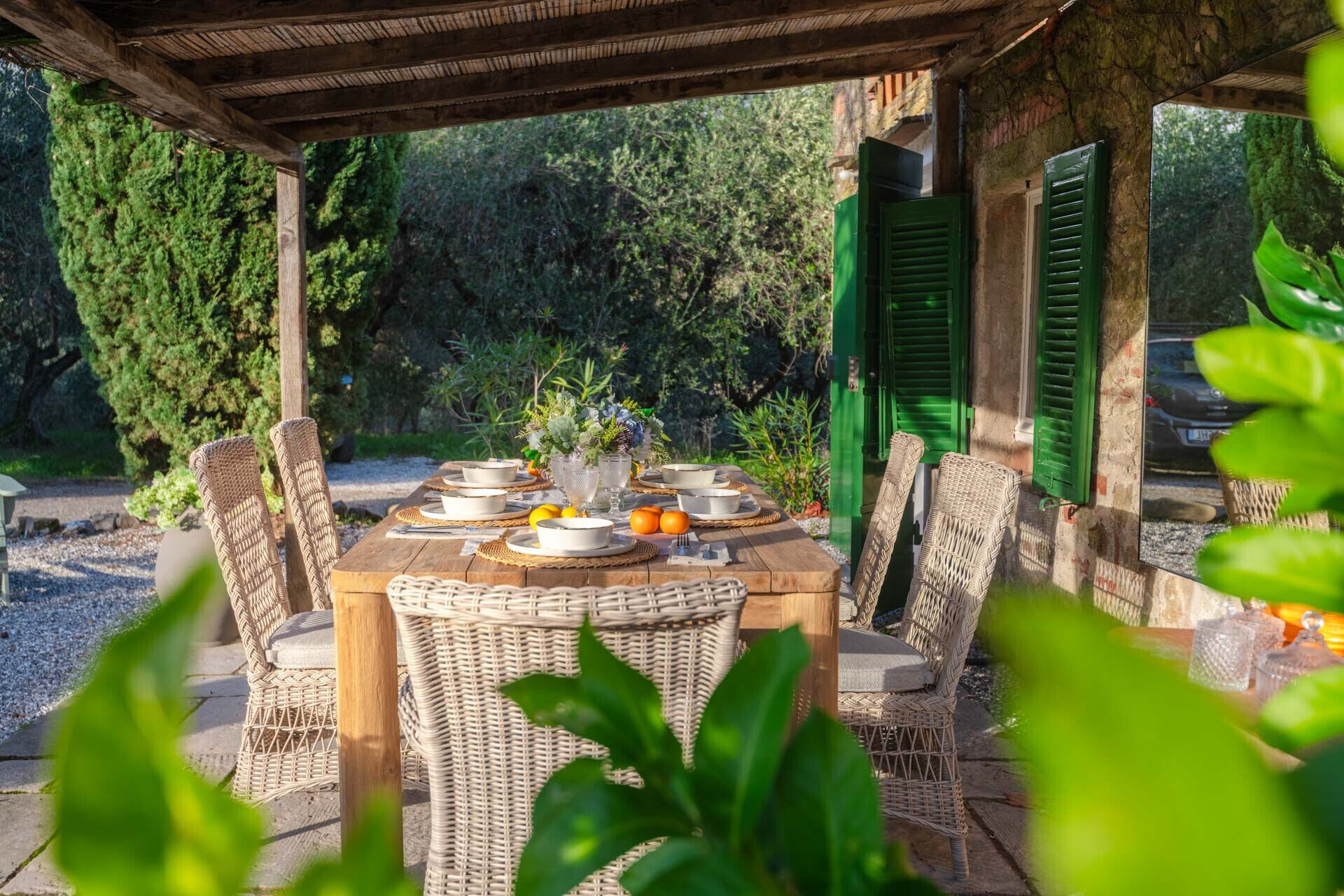 An outdoor dining area with wicker chairs and a wooden table set for a meal, surrounded by greenery and a stone wall.