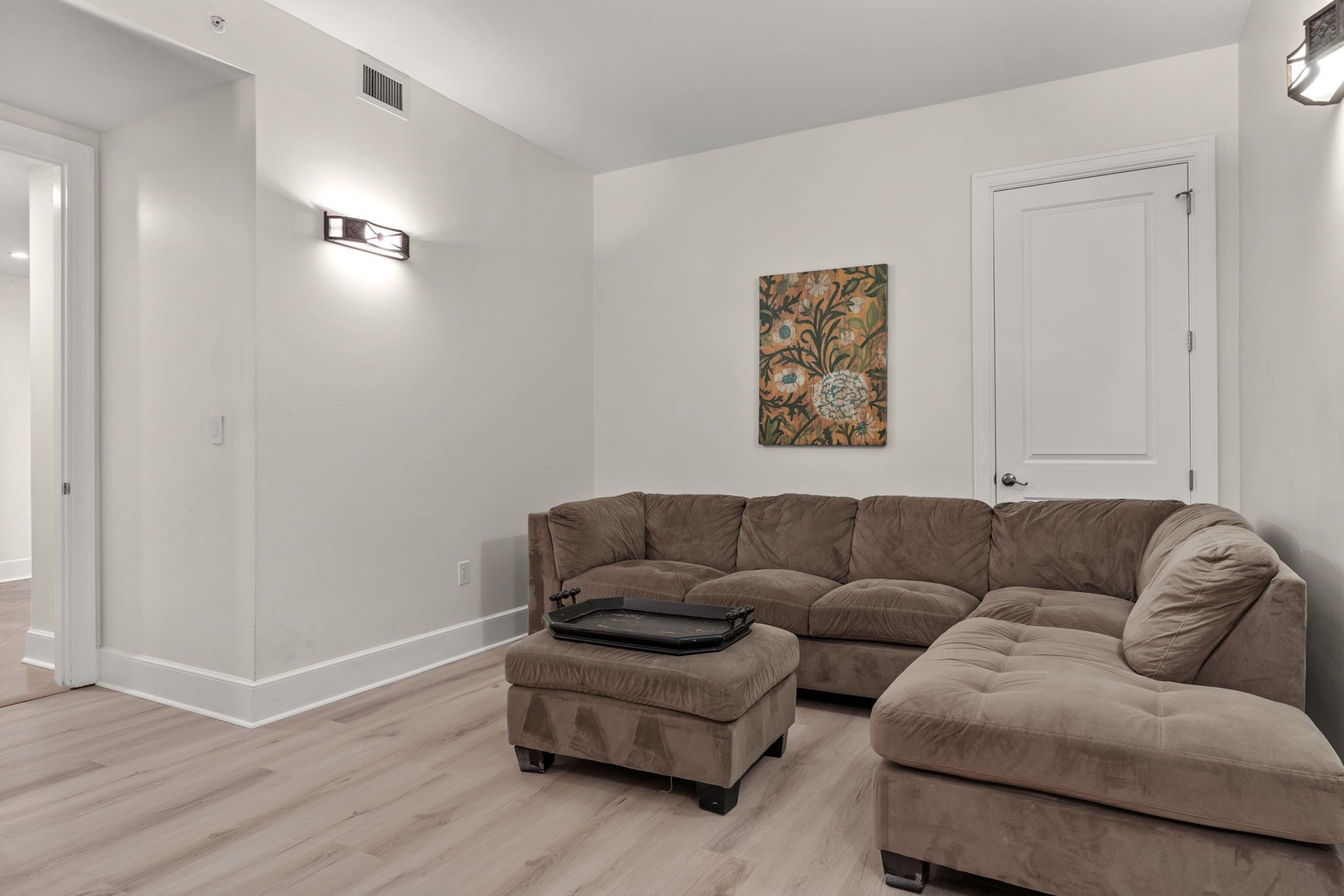 Un salon avec un canapé d’angle, un pouf et un tableau accroché au mur.
