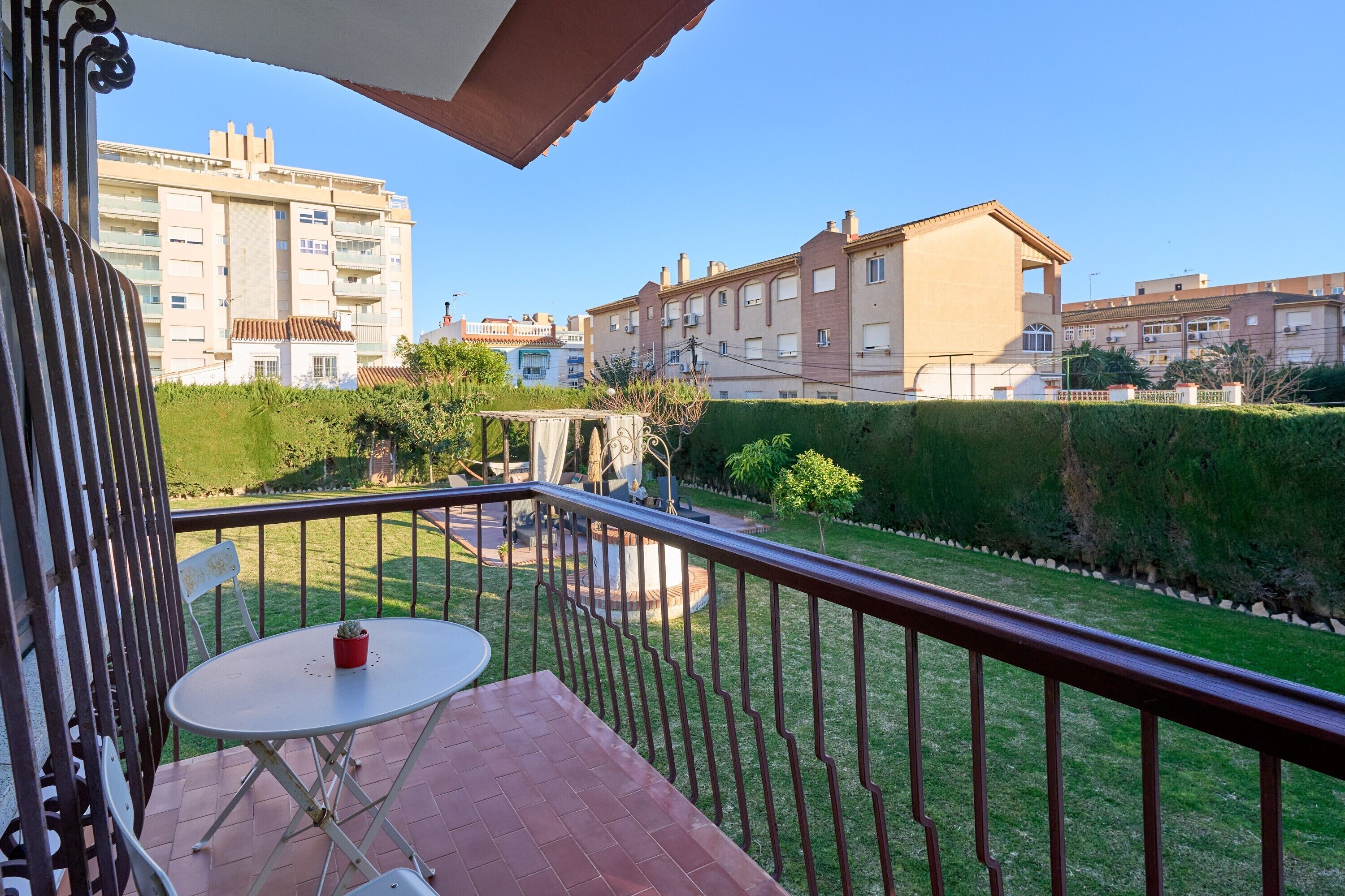 A balcony with a table and chairs, a view of a well-maintained garden, and residential buildings in the background.