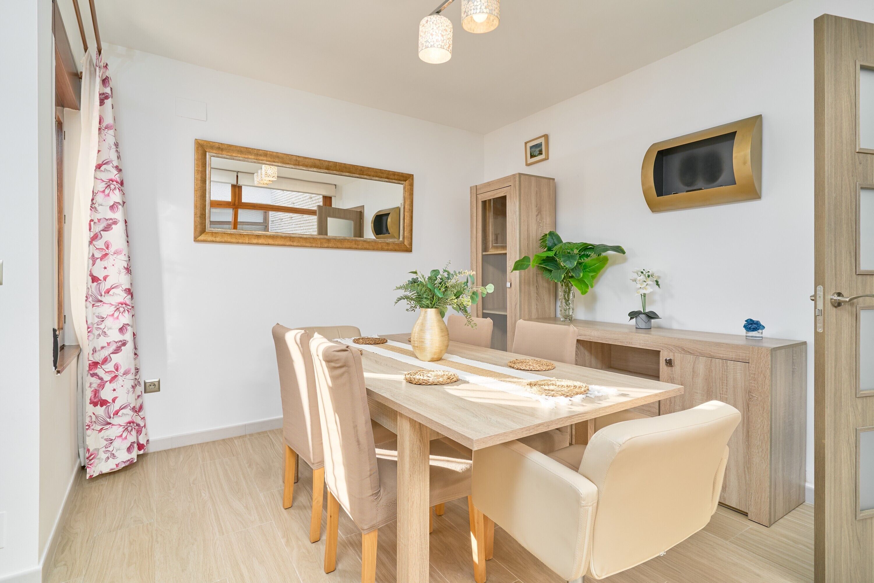 A dining area with a wooden table, beige chairs, a vase with flowers, a mirror, and a cabinet with a plant.