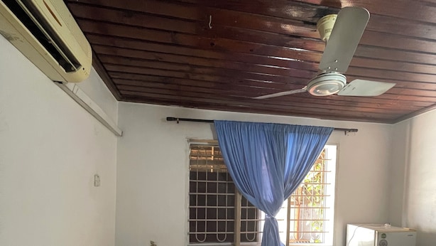A room with a bed, a ceiling fan, a window with bars, and a small table with a television. - Karibu nest hostel (Dar es Salaam)