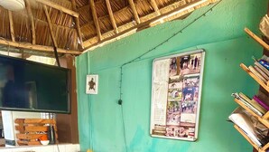 A room with a thatched roof, a green wall, a plaid sofa with a yellow pillow, a red plaid bedspread, a wooden bench, a bulletin board, and a small stool. - Karibu nest hostel (Dar es Salaam)
